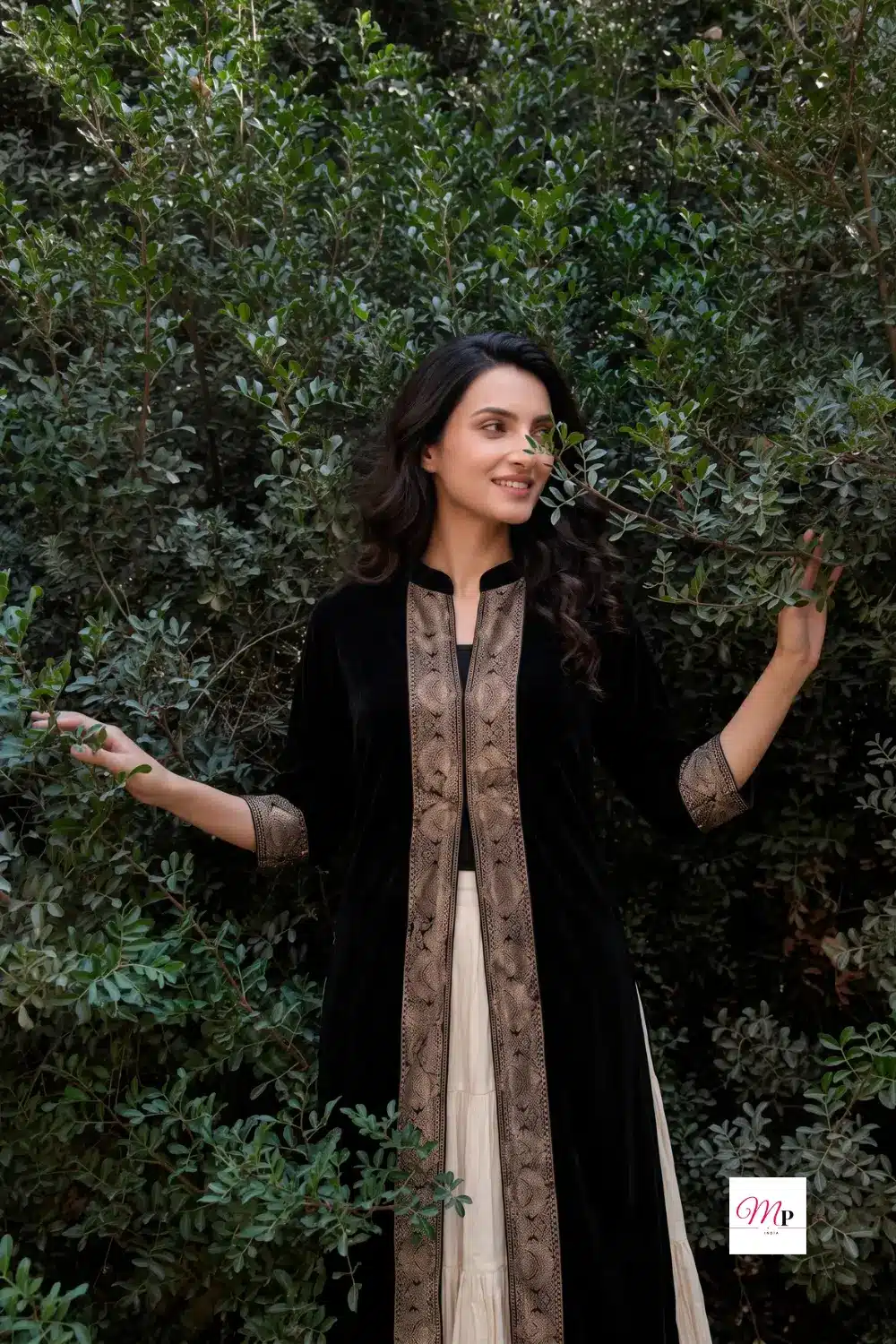 Full length portrait of black velvet anarkali with cream lehenga against green garden foliage