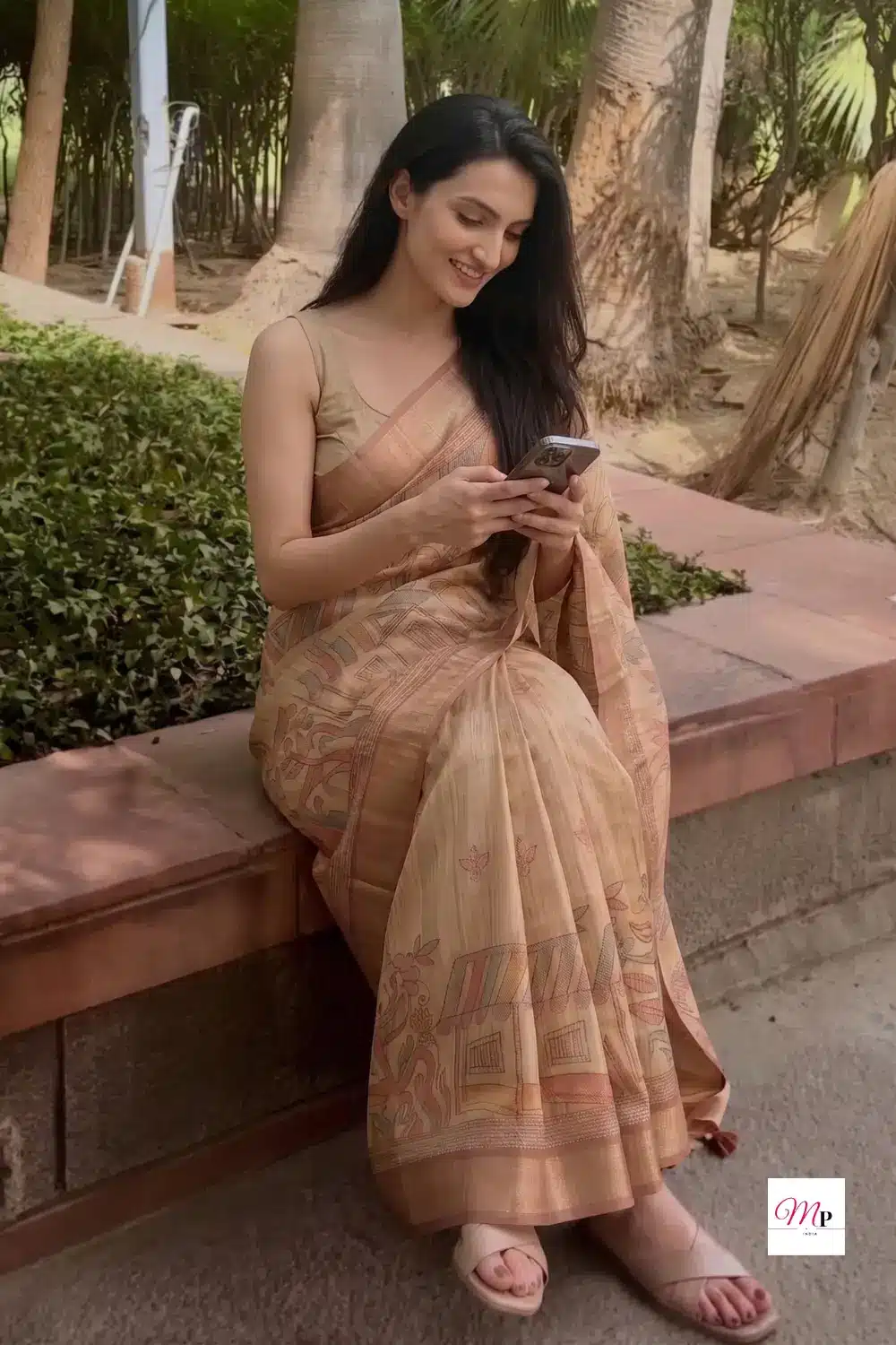 Woman wearing ethnic silk blend saree with warli motifs sitting casually and checking phone in park
