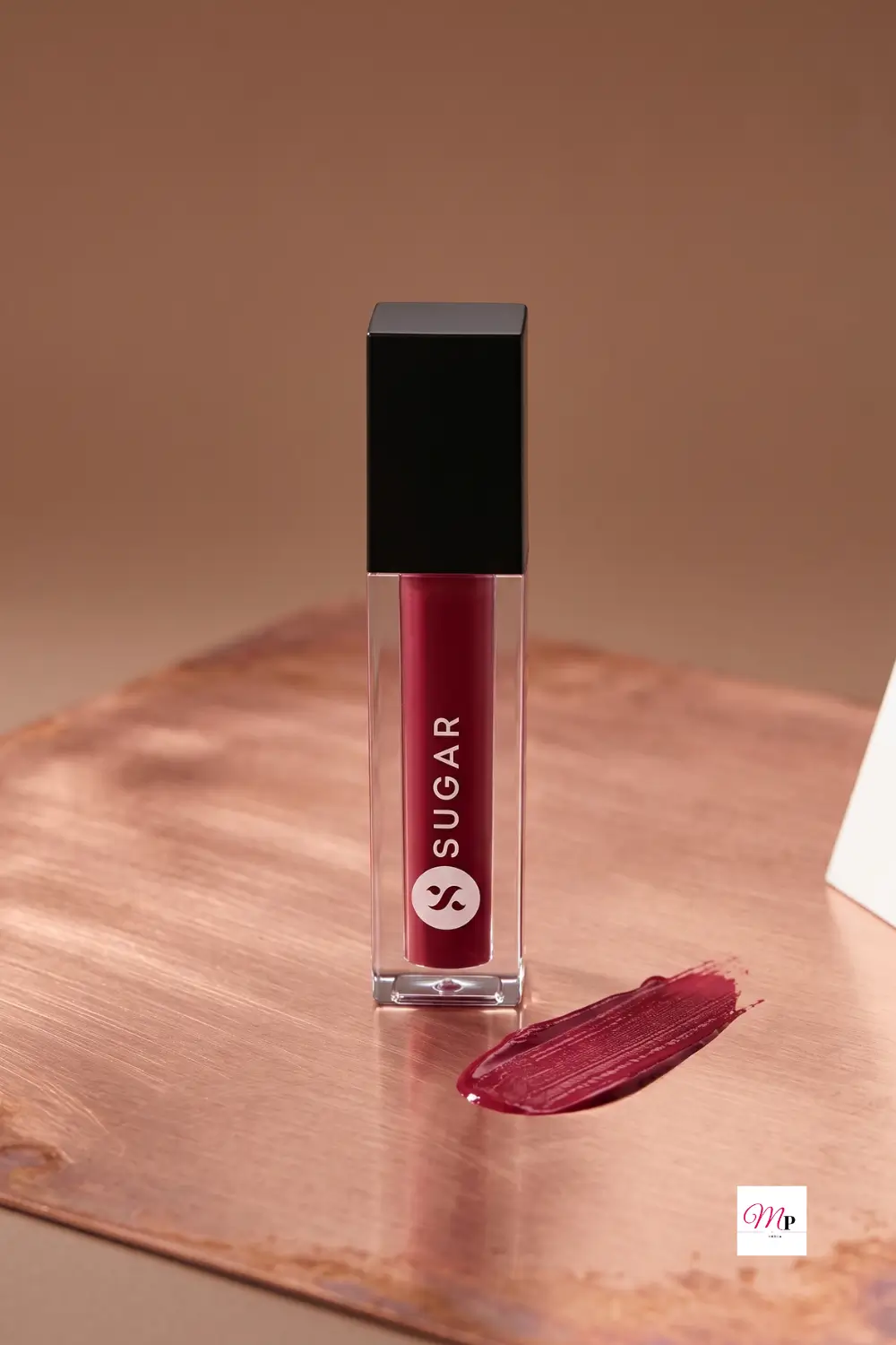 SUGAR Smudge Me Not Mini Drop Dead Red lipstick tube and burgundy swatch on warm copper wooden surface showing matte finish texture