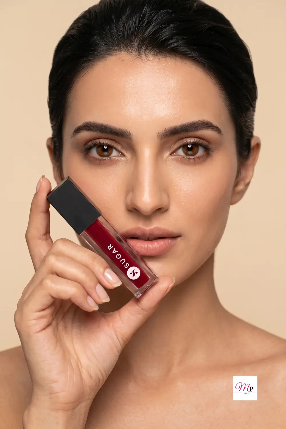 Model holding SUGAR Smudge Me Not Mini Drop Dead Red lipstick near face with natural makeup look on light beige background