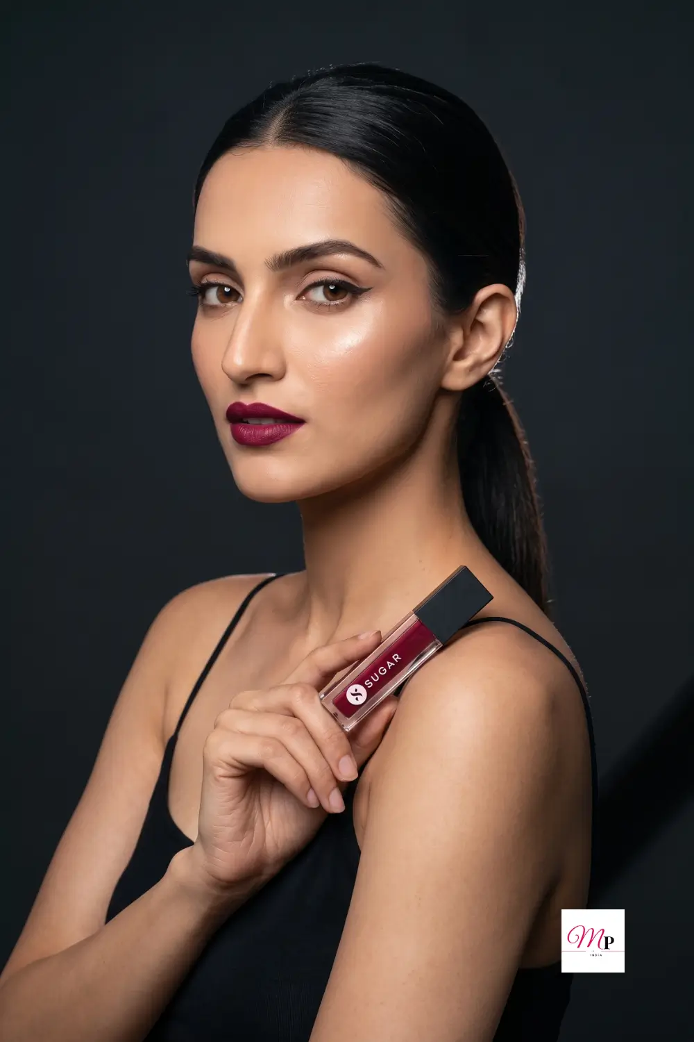 Model wearing SUGAR Smudge Me Not Mini Drop Dead Red deep red matte lipstick holding the tube near shoulder against dark background