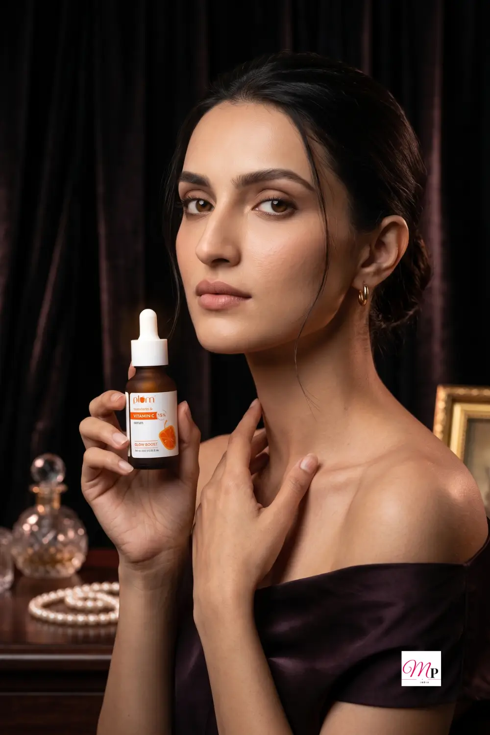 Model in off-shoulder evening look holding Plum 15% Vitamin C serum at burgundy velvet vanity with perfume bottle and pearls