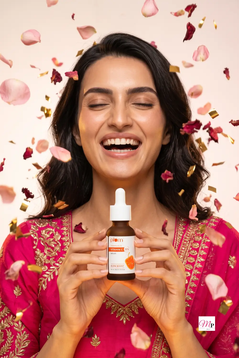 Laughing woman in festive pink embroidered outfit with falling rose petals and gold confetti holding Plum Vitamin C serum — shaadi season glow serum for Indian skin