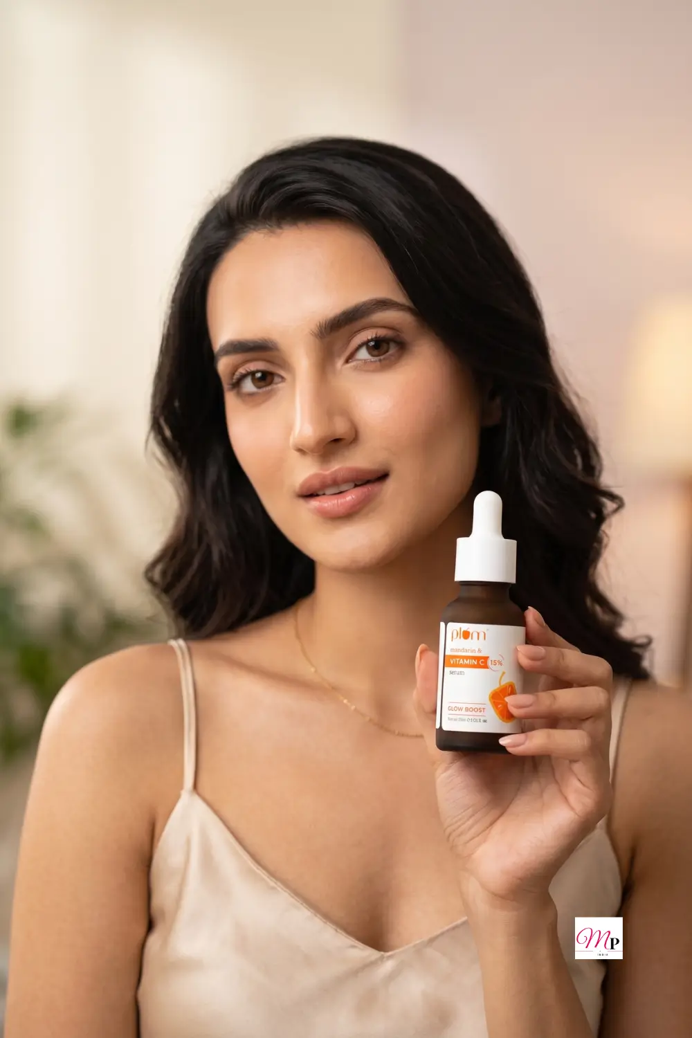 Woman in champagne silk camisole holding Plum Vitamin C glow serum near her shoulder in soft warm indoor light — serum for glowing skin