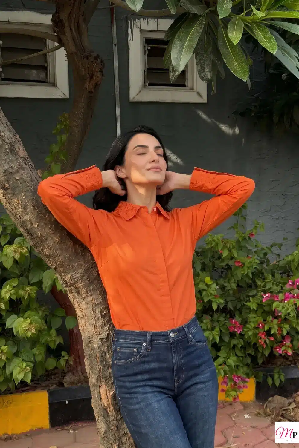 Red formal shirt styled casually with blue jeans, model leaning against tree in sunlight